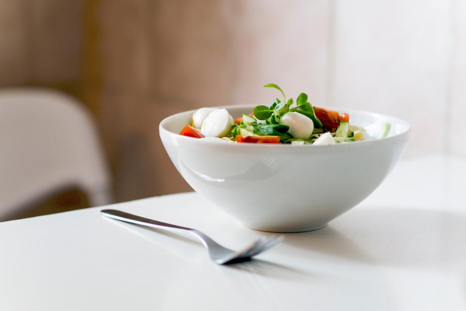 Ensalada con hojas verdes, tomate y mozzarella en un tazón blanco, sobre una mesa blanca con un tenedor